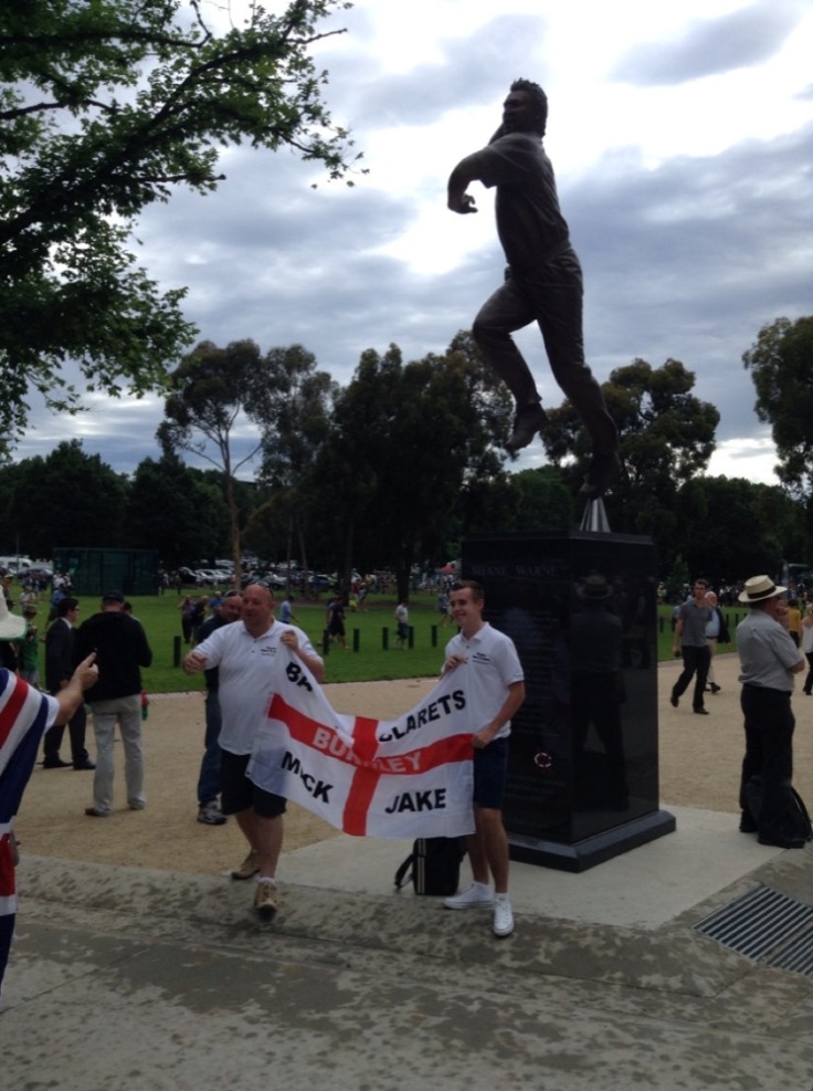 Two England fans hold up their flag in front of a statue of Shane Warne, in his bowling action, up on a plinth