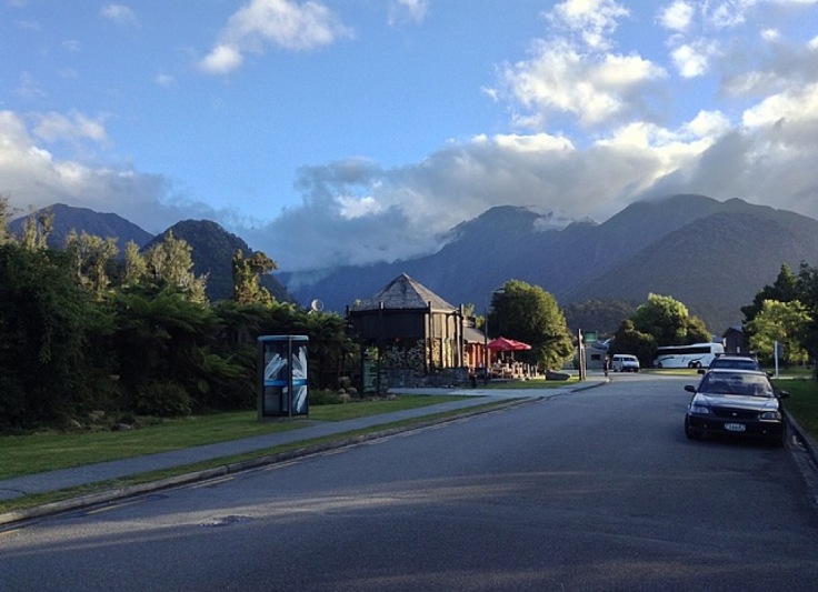 A wide street, with a phone box and car, the mountains behind look dramatic, as clouds envelop the tips