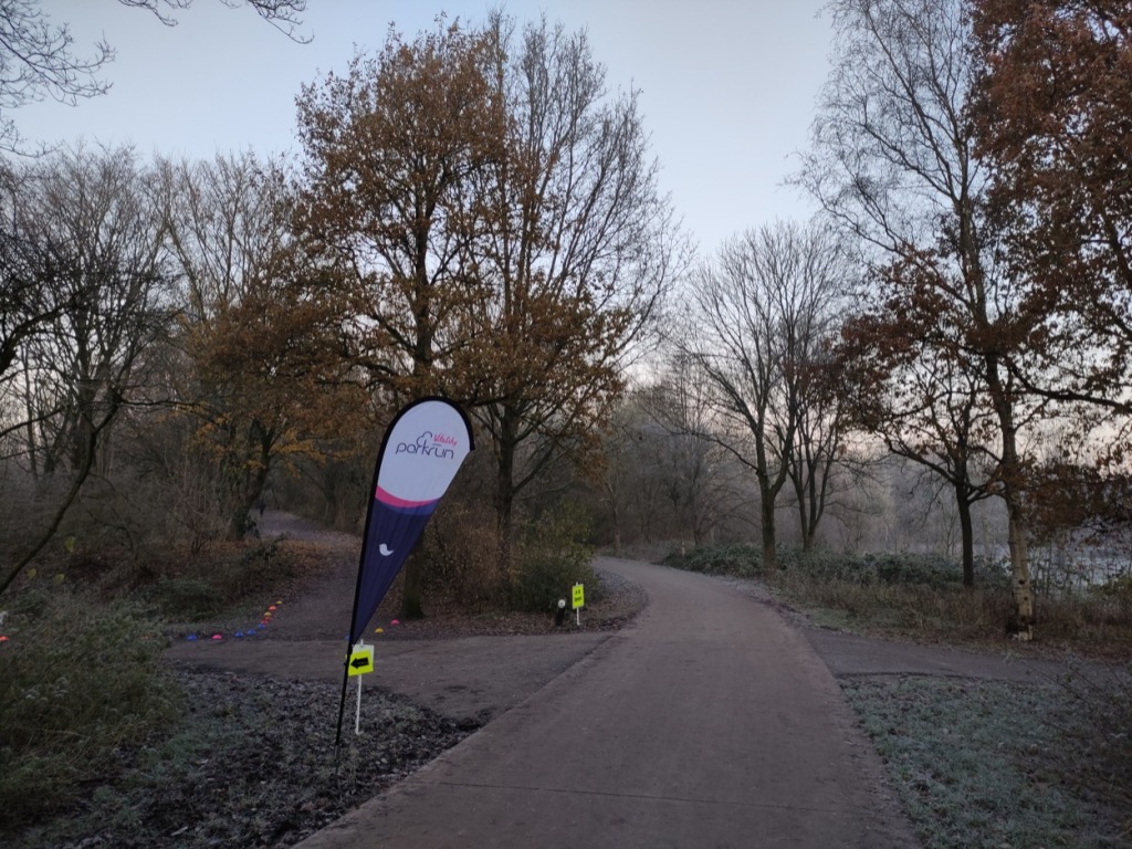 The parkrun feather flag flying next to tarmacced paths, lined with orange-leaved trees.