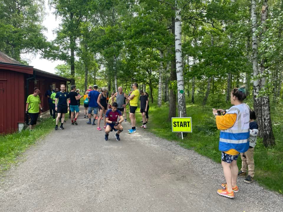 27 runners and walkers stand on the start line with a wood cabin on one side and trees on the right of a gravelly path.