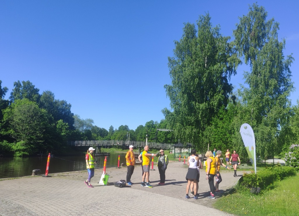A bridge over the water next to a paved area from where the parkrun starts and finishes. Several people milling around in Hi-Viz and parkrun vests.