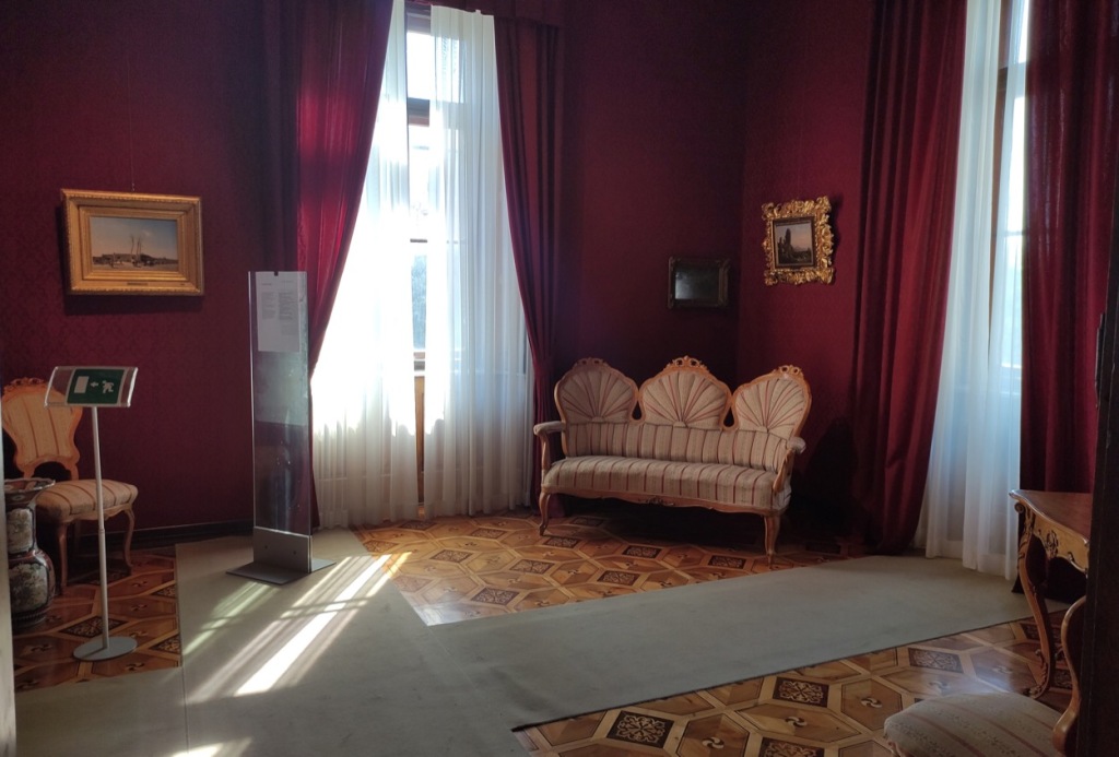 A tall room with purple walls and curtains. In the corner is a small screen which is supplied with a view of the square outside by a system of mirrors.