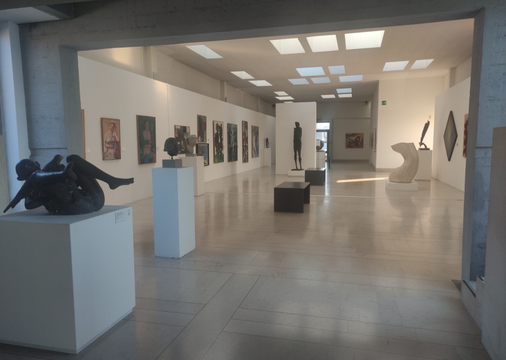 A medium-sized open gallery has modern art on the walls and sculptures on different plinths around the room. Lots of skylights allow natural light into the gallery