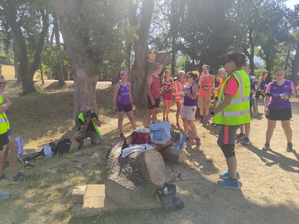 People in purple, red, hi-viz and other colours stand around talking in groups after the event has finished. Bags rest on a bench made from a cut-down tree stump.